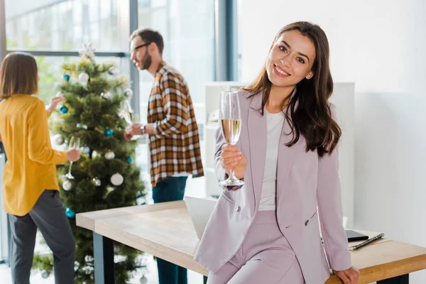 Messa a fuoco selettiva di attraente donna d'affari sorridente mentre tiene bicchiere di champagne vicino a colleghi e albero di Natale — Foto stock