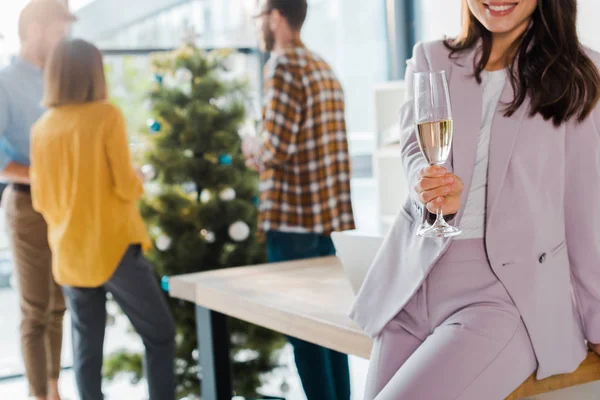 Cropped view of businesswoman holding champagne glass near coworkers and christmas tree — Stock Photo