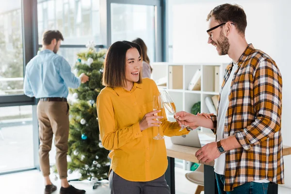 Selective focus of happy businessman and asian businesswoman toasting champagne glasses near colleagues and christmas tree — Stock Photo