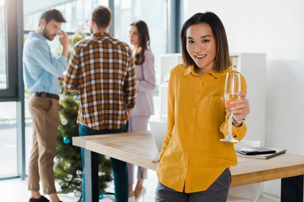 Foco seletivo de mulher asiática feliz segurando copo de champanhe perto de colegas de trabalho e árvore de natal no escritório — Fotografia de Stock