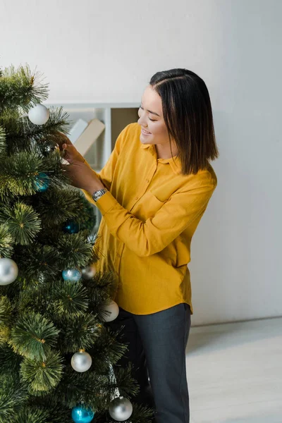 Cheerful asian woman decorating christmas tree in office — Stock Photo