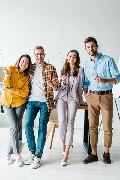 Cheerful businessmen and multicultural businesswomen holding shiny sparklers in office — Stock Photo