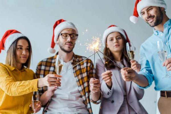 Selective focus of cheerful businessmen and multicultural businesswomen holding sparklers and champagne glasses in office — Stock Photo