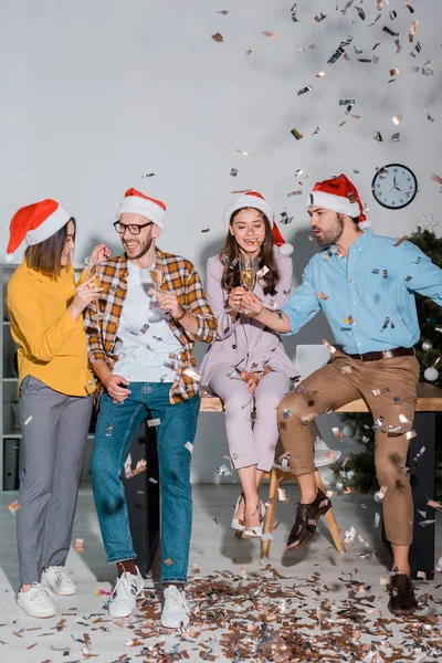 Cheerful businessmen and multicultural businesswomen in santa hats with champagne glasses near falling confetti — Stock Photo
