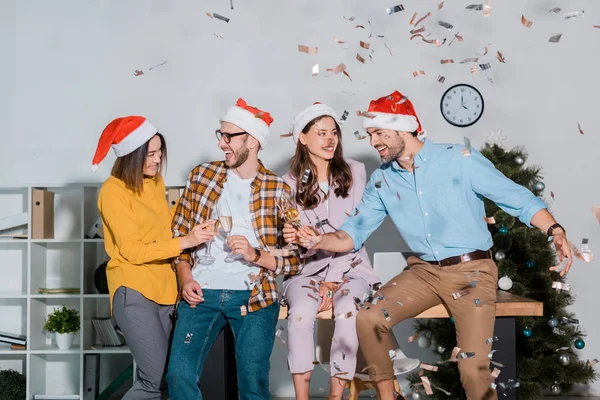 Cheerful businessmen and multicultural businesswomen in santa hats toasting champagne glasses near falling confetti — Stock Photo