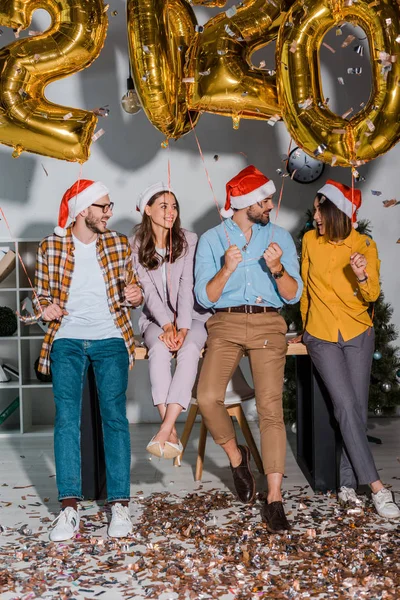 Hombres de negocios felices y mujeres de negocios multiculturales en sombreros de santa cerca de confeti y globos con números - foto de stock