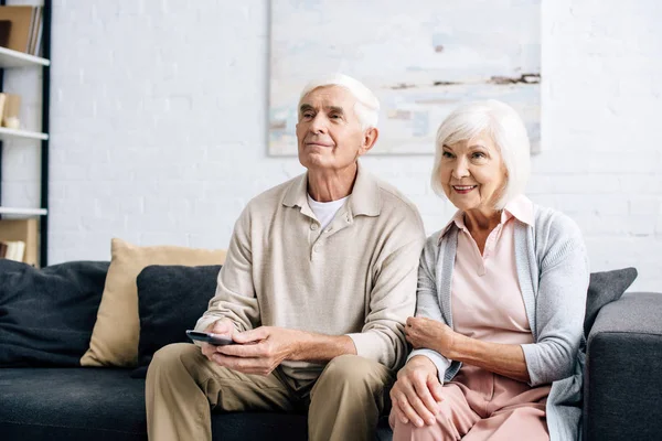 Sonrientes marido y mujer viendo la televisión y sentado en el sofá en el apartamento - foto de stock