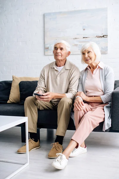 Улыбающиеся муж и жена смотрят телевизор и сидят на диване в квартире — стоковое фото