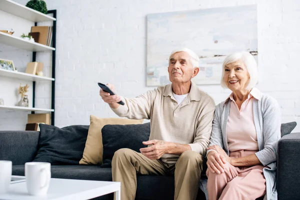 Sonrientes marido y mujer viendo la televisión y sentado en el sofá en el apartamento - foto de stock