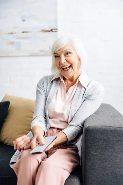 Lächelnde Seniorin sitzt auf Sofa und schaut in Wohnung fern — Stockfoto