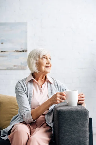 Mujer mayor sonriente sentada en el sofá y sosteniendo la taza en el apartamento — Stock Photo