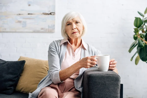 Seniorin sitzt auf Sofa und hält Tasse in Wohnung — Stockfoto