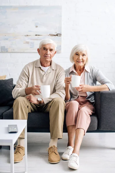 Sonrientes marido y mujer sosteniendo copas y sentado en el sofá en el apartamento - foto de stock