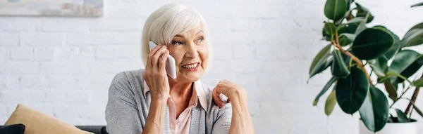 Панорамный снимок улыбающейся пожилой женщины, разговаривающей на смартфоне в квартире — стоковое фото