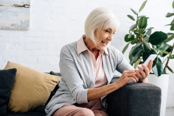 Donna anziana sorridente seduta sul divano e utilizzando smartphone in appartamento — Foto stock