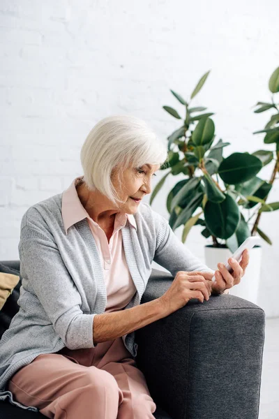 Femme âgée assise sur le canapé et utilisant un smartphone dans l'appartement — Photo de stock