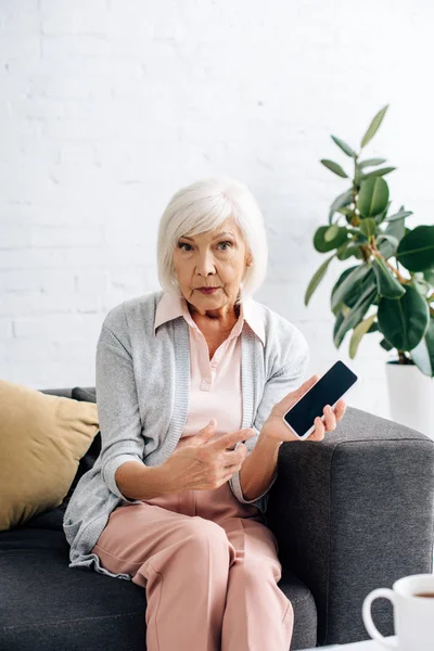 Donna anziana scioccata seduta sul divano e che punta con il dito verso smartphone in appartamento — Foto stock
