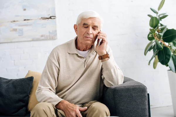 Homem sênior sentado no sofá e falando no smartphone no apartamento — Fotografia de Stock