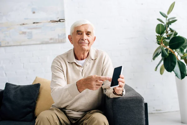 Smiling senior man sitting on sofa and pointing with finger at smartphone in apartment — Stock Photo