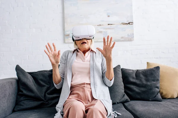 Mulher sênior chocado com fone de ouvido realidade virtual sentado no sofá no apartamento — Fotografia de Stock