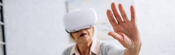 Панорамный снимок пожилой женщины в гарнитуре виртуальной реальности с протянутой рукой в квартире — стоковое фото
