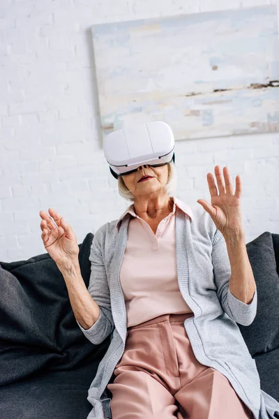 Mujer mayor en auriculares de realidad virtual con las manos extendidas en el apartamento - foto de stock