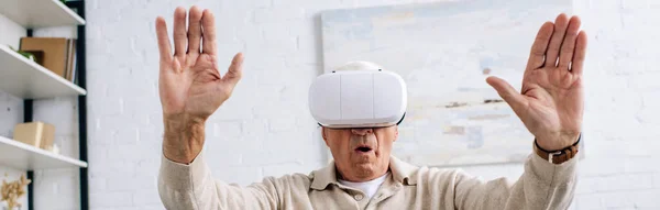 Tiro panorâmico de homem sênior em fone de ouvido realidade virtual com as mãos estendidas no apartamento — Fotografia de Stock