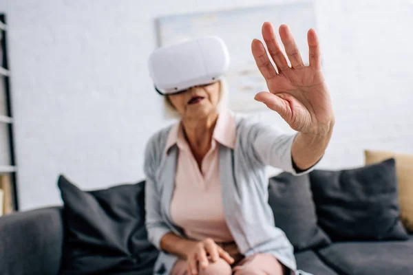 Enfoque selectivo de la mujer mayor en auriculares de realidad virtual con la mano extendida en el apartamento - foto de stock