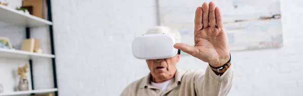 Tiro panorâmico de homem sênior em fone de ouvido realidade virtual com mão estendida no apartamento — Fotografia de Stock