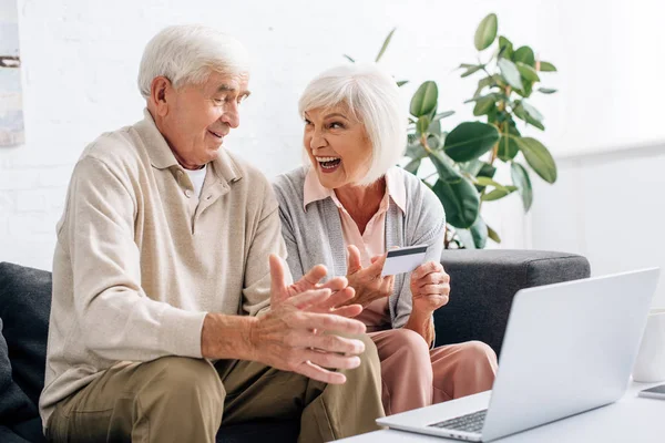 Sorrindo marido e mulher usando laptop e segurando cartão de crédito no apartamento — Fotografia de Stock