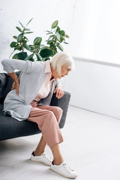Mulher sênior tendo dor nas costas e sentado no sofá no apartamento — Fotografia de Stock