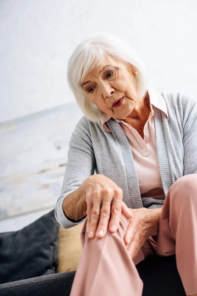 Vista angolo basso della donna anziana che ha l'artrite del ginocchio e seduta sul divano in appartamento — Foto stock