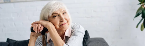 Panoramic shot of smiling senior woman with wooden cane looking at camera in apartment — Stock Photo