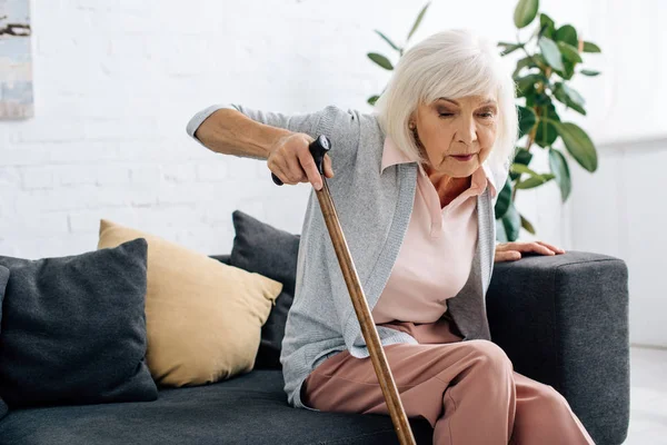 Mujer mayor con bastón de madera sentado en el sofá en el apartamento — Stock Photo