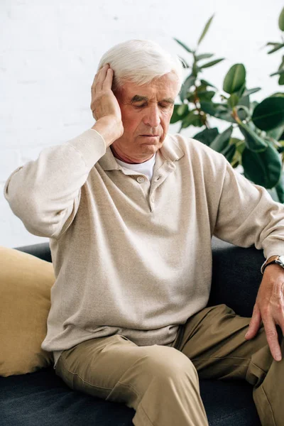 Hombre mayor sentado en el sofá y con dolor de cabeza en el apartamento - foto de stock