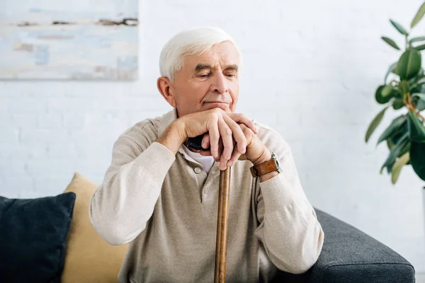 Senior man sitting on sofa and holding wooden cane in apartment — Stock Photo