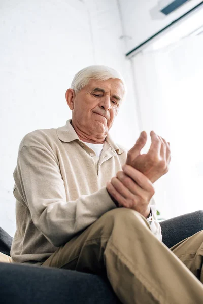 Low angle view of senior man sitting on sofa and having pain in hand in apartment — Stock Photo