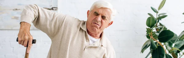 Panoramic shot of senior man having pain and holding wooden cane in apartment — Stock Photo