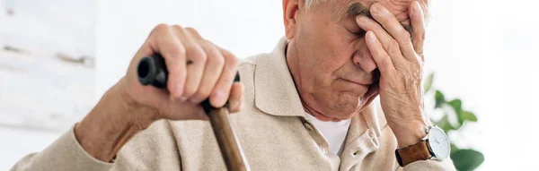 Plano panorámico del hombre mayor que tiene dolor de cabeza y la celebración de caña de madera en el apartamento - foto de stock