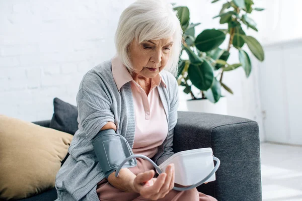 Пожилая женщина сидит на диване и измеряет артериальное давление в квартире — стоковое фото