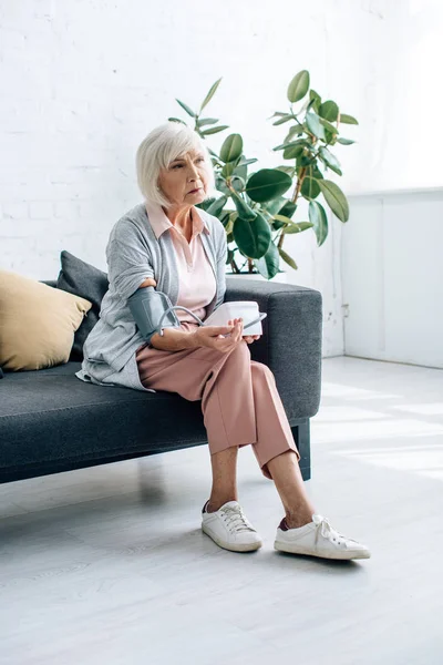 Пожилая женщина сидит на диване и измеряет артериальное давление в квартире — стоковое фото