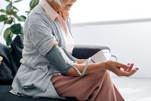 Vista ritagliata di donna anziana seduta sul divano e misurazione della pressione sanguigna in appartamento — Foto stock