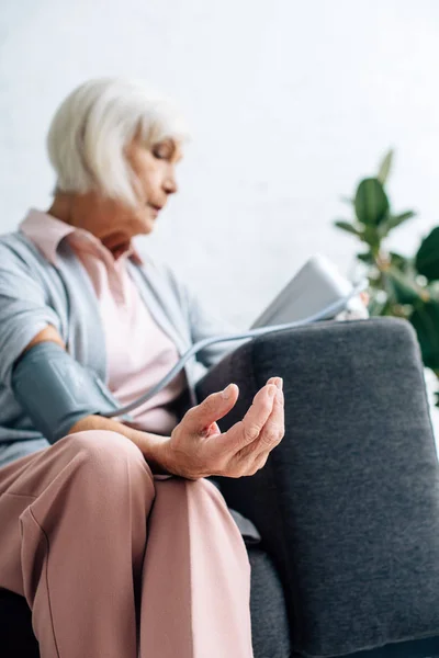 Селективное внимание пожилой женщины, сидящей на диване и измеряющей артериальное давление в квартире — стоковое фото