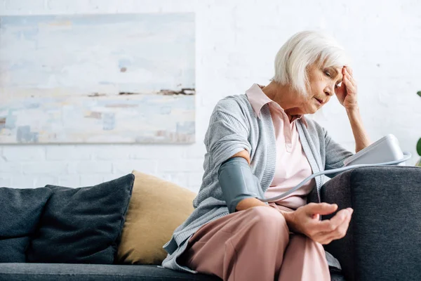 Старша жінка сидить на дивані і вимірює кров'яний тиск в квартирі — стокове фото
