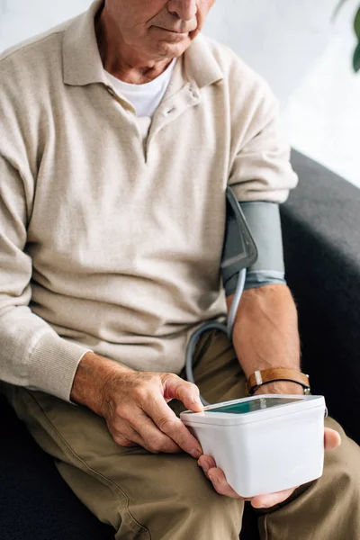 Cropped view of senior man measuring blood pressure and sitting on sofa in apartment — Stock Photo