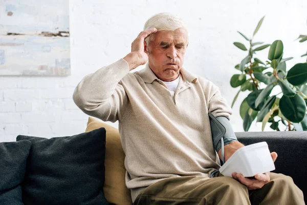 Shocked senior man measuring blood pressure and sitting on sofa in apartment — Stock Photo