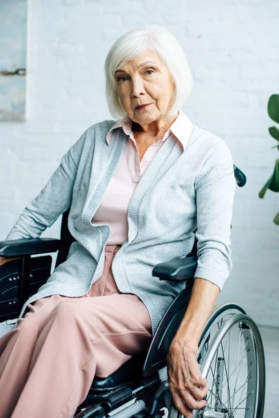 Старша жінка сидить у інвалідному кріслі, дивлячись на камеру в квартирі — стокове фото