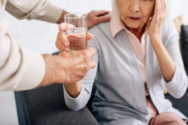 Vista ritagliata del marito che dà un bicchiere d'acqua alla moglie con mal di testa in appartamento — Foto stock