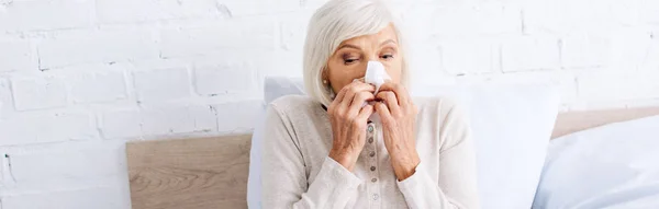 Panoramic shot of ill senior woman sneezing and holding napkin in apartment — Stock Photo