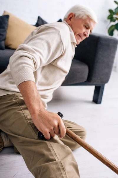 Selective focus of senior man holding wooden cane and feeling pain in apartment — Stock Photo